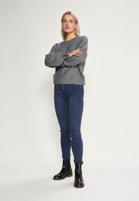 Šedý pletený svetr s volným střihem a balónovými rukávy, v kombinaci s tmavě modrými skinny džíny a lesklými černými kotníkovými botami.