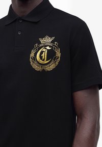 Μαύρο πόλο με χρυσό έμβλημα Just Cavalli Creative Department στο αριστερό μέρος του στήθους, φορεμένο από άτομο με σκούρο δέρμα.