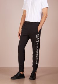 Čierne teplákové nohavice s elastickým pásom. Biele logo "EA7 EMPORIO ARMANI" vytlačené po ľavej nohe. Nosené s čiernymi teniskami.