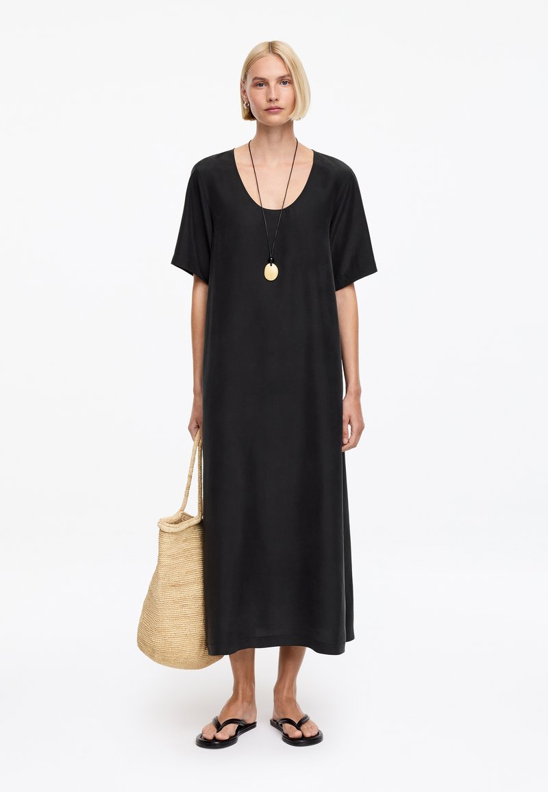 Femme portant une robe midi noire ample, des sandales noires, un collier pendentif rond, et tenant un sac fourre-tout en paille tressée.