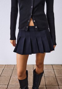 Cardigan nero a coste con bottoni a pressione, abbinato a una mini gonna nera plissettata. Stivali neri al ginocchio completano il look.