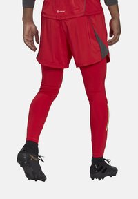 Rode sportieve shorts over rode leggings, met een gladde textuur, contrasterende grijze accenten en zwarte voetbalschoenen met gouden details.