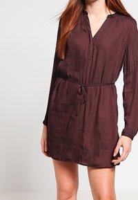 Kobieta w długiej, ciemnobrązowej sukience z paskiem, z dekoltem w kształcie litery V, stojąca na tle jednolitego koloru.