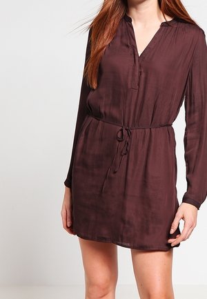 Vrouw in een lange mouw, donkerbruin, met een riem om de taille, jurk met V-hals, staande tegen een effen achtergrond.