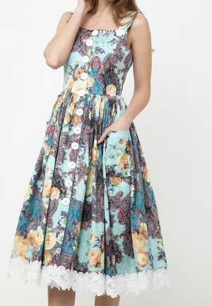 Frau trägt ärmelloses, knöpfbares Blumenkleid mit aufgesetzten Taschen und weißem Spitzenbesatz am Saum, die rechte Hand in der Tasche.