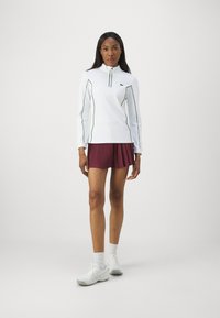 Camiseta blanca de manga larga con costuras verdes y cremallera de un cuarto, combinada con pantalones cortos burdeos con plisado y zapatillas deportivas blancas.