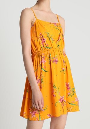 Kvinde iført en ærmeløs orange kjole med tynde stropper og pink blomsterprint, stående mod en ensfarvet lys baggrund.