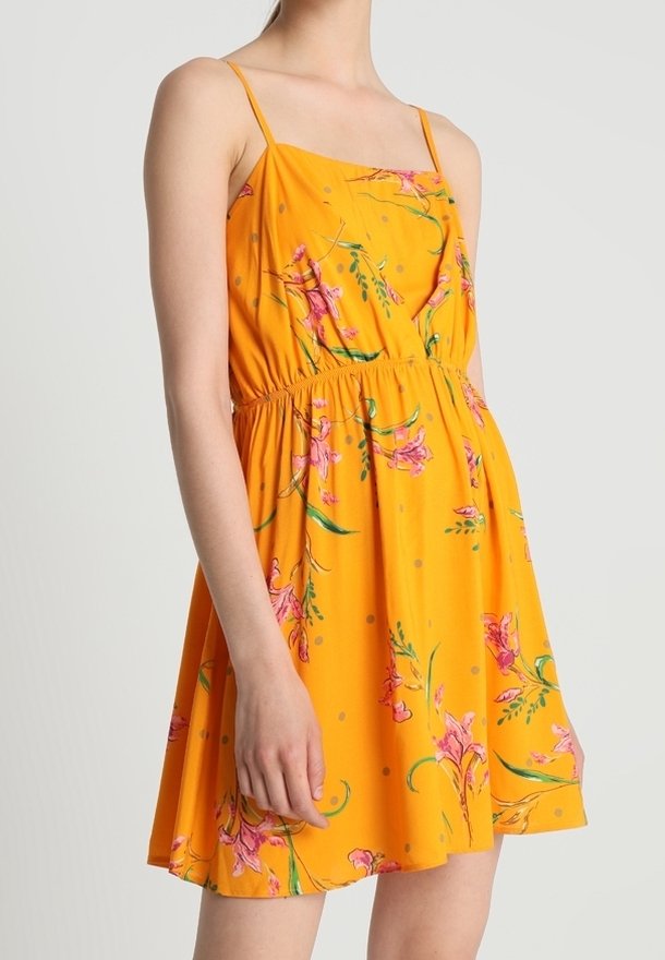 Femme portant une robe orange sans manches avec de fines bretelles et un imprimé floral rose, debout devant un fond clair uni.