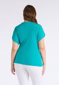 Top turchese a maniche corte con scollatura rotonda e texture liscia, caratterizzato da una leggera caduta e una cucitura lungo la schiena. Abbinato a pantaloni bianchi.