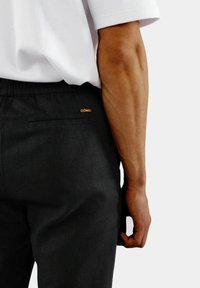 Svarte bukser med teksturert finish, som har en baklomme og et oransje logoakkent. Modellen sin arm er delvis synlig.
