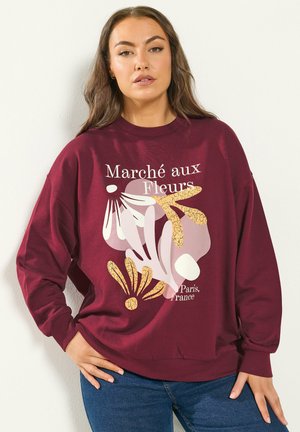 Frau trägt ein purpurrotes Sweatshirt mit abstraktem Blumenmuster und dem Text "Marché aux Fleurs, Paris, Frankreich", kombiniert mit blauer Jeans.