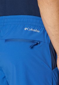 Siniset Columbia-shortsit, joissa on joustava vyötärö, sivulla vetoketjullinen tasku ja kontrastiväriset tummansiniset yksityiskohdat. Pehmeä, kevyt kangas.