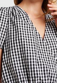 Blusa de cuadros en blanco y negro con escote en V, mangas cortas abullonadas y una tela ligera. Patrón de cuadrícula notable en toda la prenda.