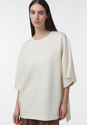 Jeune femme aux longs cheveux foncés portant un haut ample à manches courtes de couleur blanc cassé uni et un pantalon marron, sur un fond blanc.