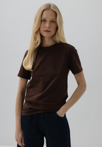 Brązowa bawełniana koszulka z krótkim rękawem i okrągłym dekoltem. Model łączy ją z ciemnoniebieskimi dżinsami. Gładka tekstura, minimalistyczny design.