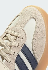 Zapatilla deportiva beige que combina materiales de ante y sintéticos, con rayas en color azul marino y cordones texturizados. Suela de goma semitranslúcida.