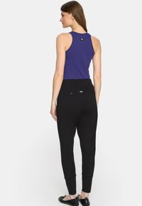 Kobieta stoi, ubrana w bezrękawnik w kolorze purpurowym i czarne spodnie z wysokim stanem z guzikami na tylnej kieszeni, zwrócona plecami na białym tle.