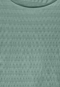 Jasnozielona teksturowana tkanina z wzorem w kratkę i subtelnymi wypukłymi liniami, z okrągłym dekoltem i gładkim wykończeniem.