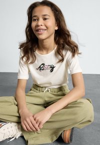 T-shirt crème à manches courtes avec un logo "Roxy" coloré. Pantalons verts olive à coupe décontractée avec une taille élastique. Baskets blanches avec des accents marron.