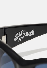 Musthave mustad prillid, millel on läbipaistev sinine toon ja millel on käe küljes hõbedane "Catfish 30th" logo, valmistatud läikivast plastmaterjalist.