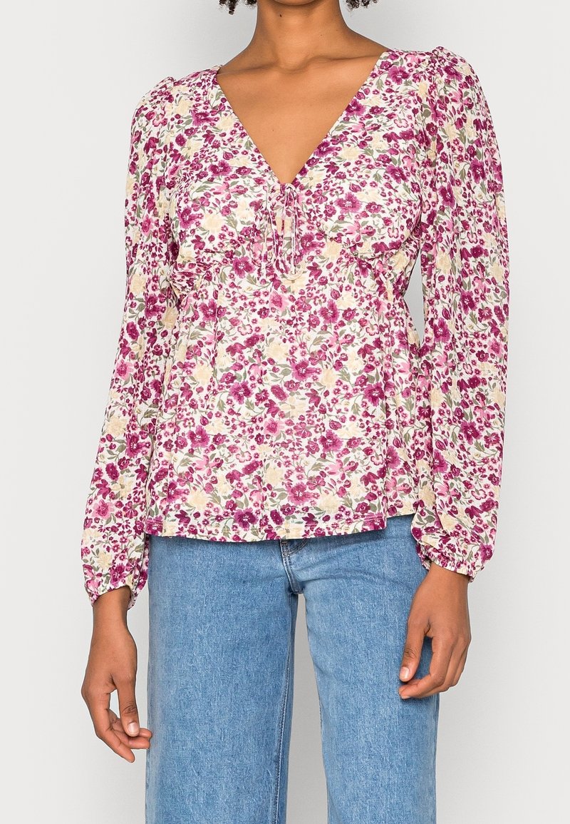 Femme portant un chemisier à manches longues à motif floral avec un col en V et un détail noué, associé à un jean en denim bleu clair.