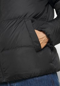 Zwarte gewatteerde jas met ritssluiting, zijzakken en een gestructureerde buitenkant. Het materiaal lijkt lichtgewicht en geïsoleerd te zijn.