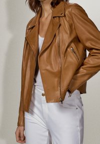 Giacca da motociclista in pelle marrone con tasche con cerniera e spalline, indossata sopra una maglietta bianca e abbinata a pantaloni bianchi. Texture liscia.