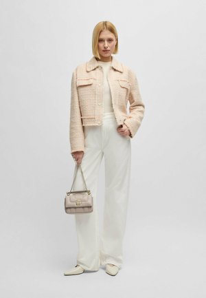 Giacca cropped in tweed rosa con quattro tasche frontali, abbinata a pantaloni larghi bianco sporco, mentre tiene una borsa grigia chiara con tracolla a catena.