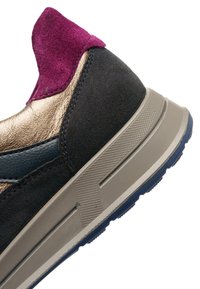 Sportschuh mit einem Obermaterial aus dunkelgrauem Wildleder, metallischen goldenen Akzenten, einer magentafarbenen Schlaufe und einer strukturierten beigen Sohle mit einem blauen Profil.