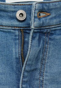 Los jeans de mezclilla azul claro presentan un cierre de cremallera y un botón de prensa de metal. La tela muestra una ligera textura con acentos de costura en tono marrón contrastante.