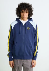 Navy zip-up hoodie with light blue shoulder panels and yellow side stripes featuring "NIKE" text. Includes a hexagonal logo on the chest.