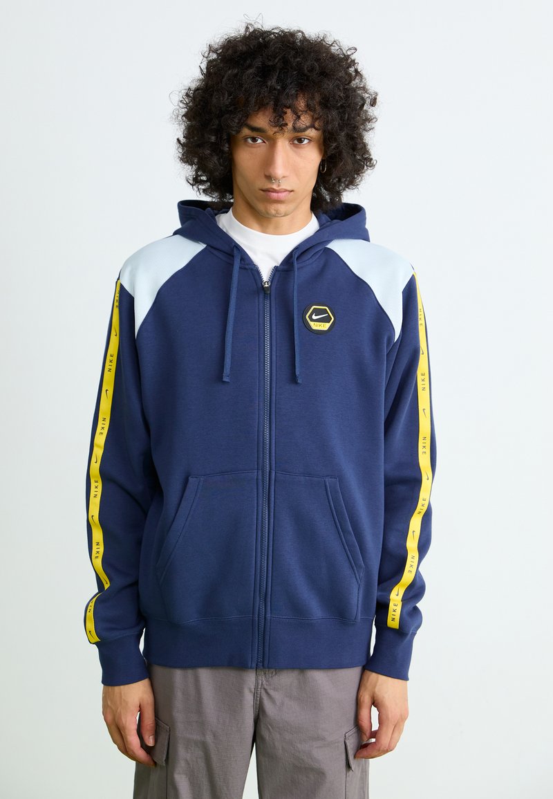 Navy zip-up hoodie with light blue shoulder panels and yellow side stripes featuring "NIKE" text. Includes a hexagonal logo on the chest.