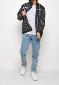 Svart randig jacka med vit text, dragkedja framtill och elastiska ärmslut, bärs över en grå t-shirt och ljusblå jeans med vita sneakers.