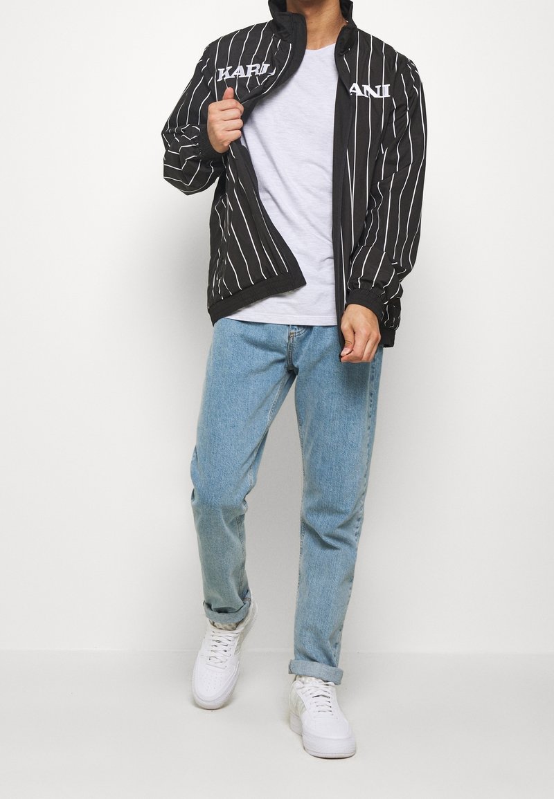 Svart randig jacka med vit text, dragkedja framtill och elastiska ärmslut, bärs över en grå t-shirt och ljusblå jeans med vita sneakers.