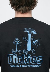 Μαύρο T-shirt με μπλε καρτούν γραφικό που απεικονίζει ένα σφυρί και μια φιγούρα από μπαστούνι, με το κείμενο "Dickies 'ALL IN A DAY'S WORK!'" από κάτω.