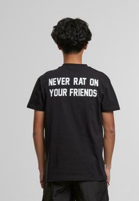 T-shirt en coton noir à manches courtes. Le dos présente un texte blanc en gras qui dit "NE JAMAIS TRAHIR SES AMIS". Encolure classique.