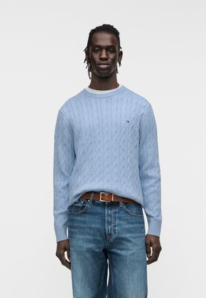 Maglione azzurro chiaro a trecce con scollo tondo, abbinato a jeans scuri e cintura marrone. Presenta polsini a coste e un piccolo dettaglio con logo.
