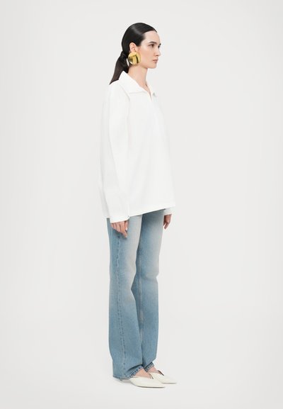 Camisa blanca de gran tamaño con cuello, jeans acampanados de color azul claro y planos blancos con punta, acentuados por grandes pendientes de aro dorados.
