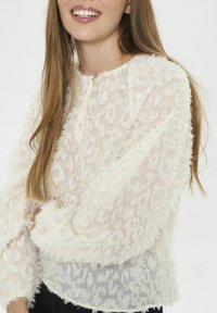 Donna che indossa una blusa bianca trasparente e strutturata a maniche lunghe con un sottile motivo geometrico, sorridendo con lunghi capelli castano chiaro.