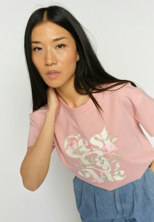 Vrouw met lang zwart haar, gekleed in een roze T-shirt met bloemenprint en blauwe broek met hoge taille, poseert met haar hand bij haar hals tegen een effen achtergrond.