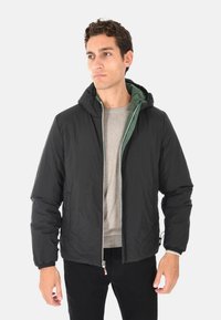 Uomo con un cappotto puffer nero con cappuccio sopra un maglione beige e pantaloni neri, che guarda leggermente di lato su uno sfondo bianco.