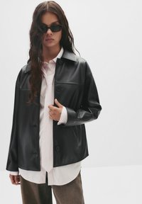 Schwarze Kunstlederjacke mit Knopfverschluss, getragen über einem hellrosa gestreiften Hemd, das eine Krawatte und hellbraune Hosen kombiniert.
