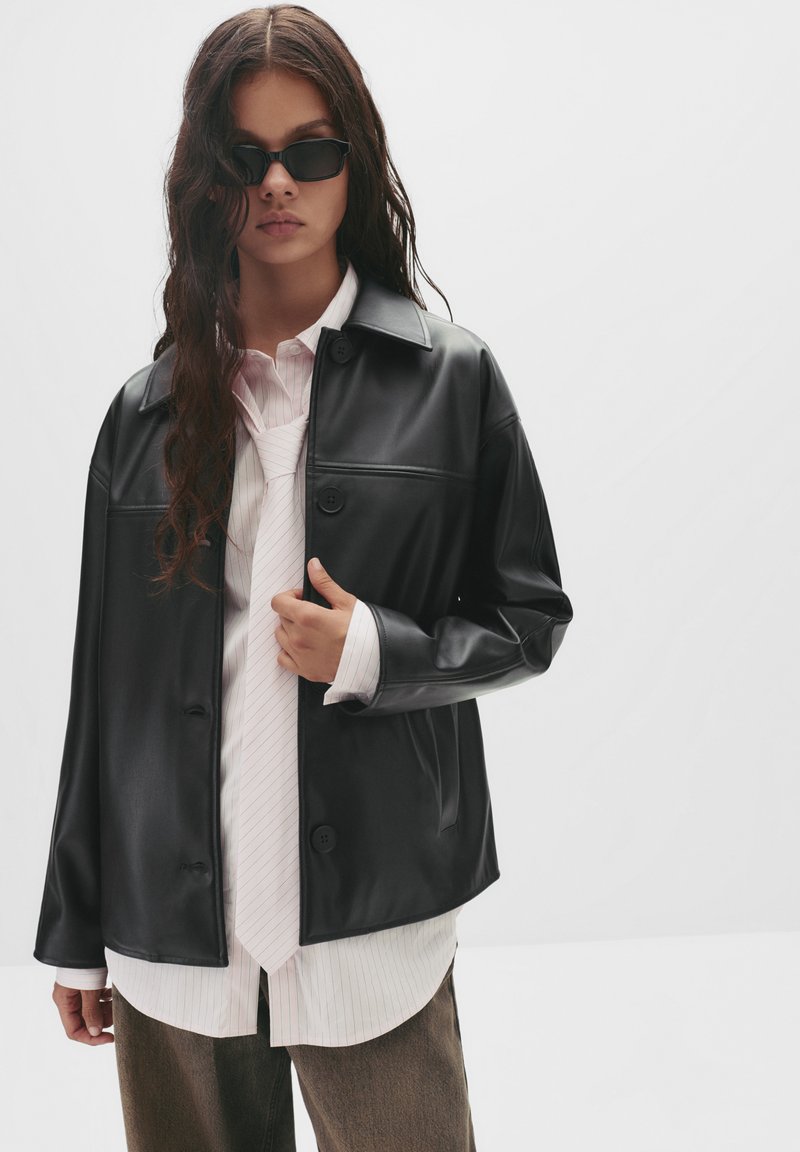 Schwarze Kunstlederjacke mit Knopfverschluss, getragen über einem hellrosa gestreiften Hemd, das eine Krawatte und hellbraune Hosen kombiniert.