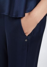 Granatowe satynowe spodnie posiadają boczną kieszeń z małym srebrnym akcentem przy otwarciu. Tkanina ma gładką, błyszczącą teksturę.