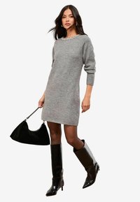 Rochie tricotată gri, cu un decupaj decorat cu mărgele la gât, purtată cu botine din piele neagră până la genunchi și o geantă neagră. Design simplu, ajustat.