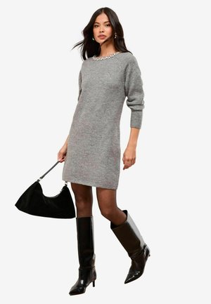 Graues Strickkleid mit einer bestickten Halslinie, getragen mit kniehohen schwarzen Lederstiefeln und einer schwarzen Handtasche. Schlichtes, tailliertes Design.