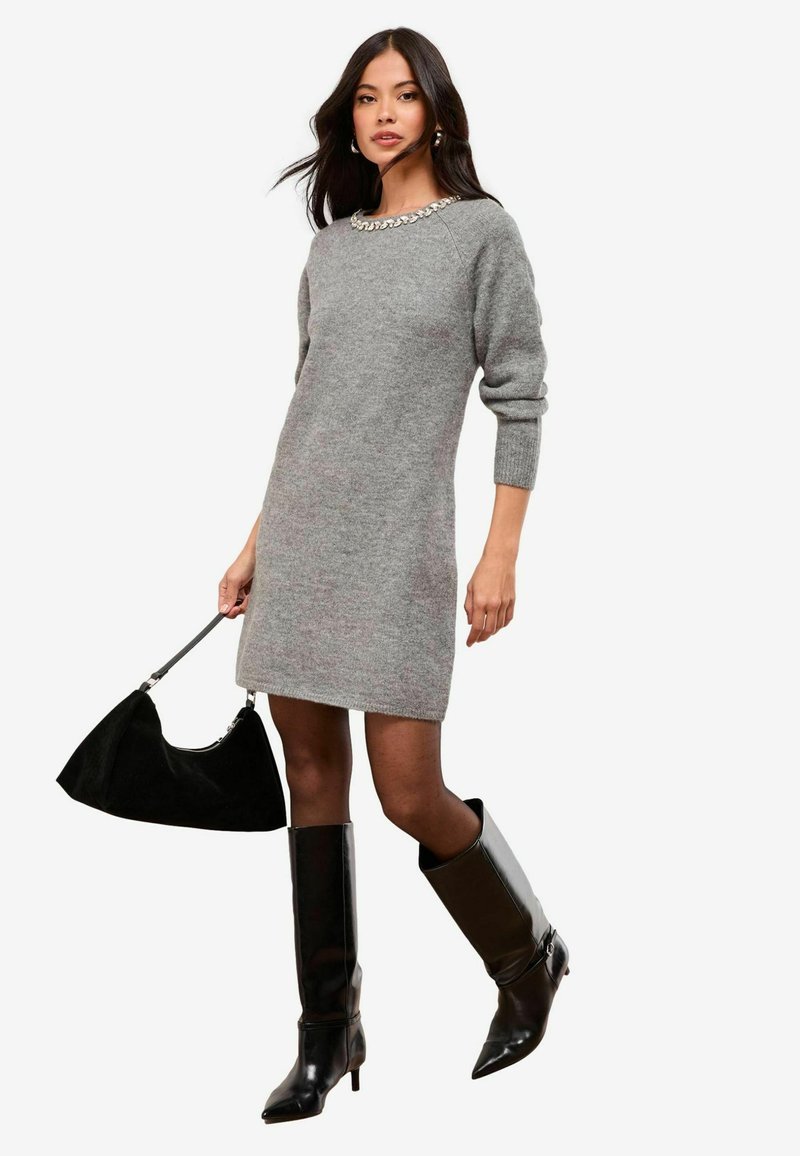 Rochie tricotată gri, cu un decupaj decorat cu mărgele la gât, purtată cu botine din piele neagră până la genunchi și o geantă neagră. Design simplu, ajustat.