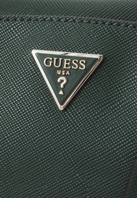 Logo triangulaire noir et argent Guess USA avec un point d'interrogation sur une surface en cuir vert foncé texturée.
