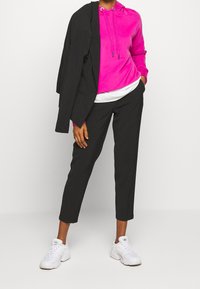 Felpa fucsia con cappuccio e stringhe sotto un blazer nero, abbinata a pantaloni neri alla caviglia e sneakers bianche da ginnastica. Tessuto morbido e design fitted.