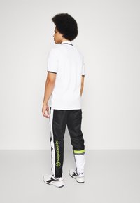 Vit polo skjorta med svarta detaljer, i kombination med svarta träningsbyxor med vita ränder och gula accenter, märkt med "Sergio Tacchini."